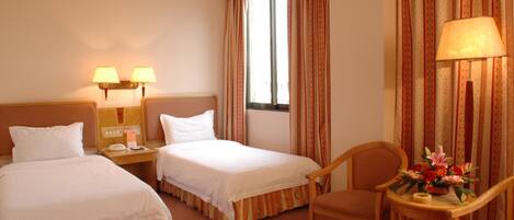 A hotel room with two beds, a desk with a lamp, a chair, and a window with curtains.
