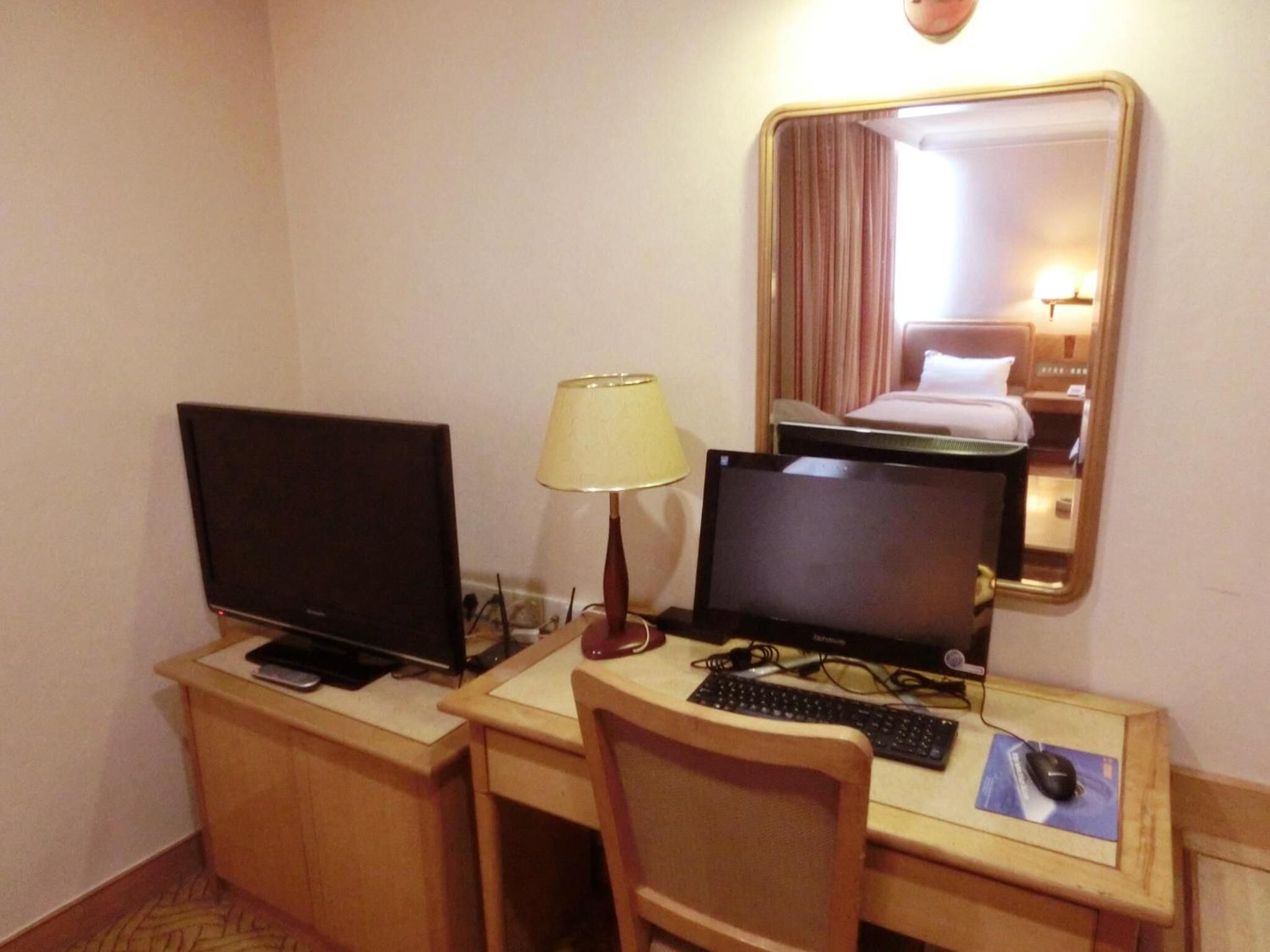 A hotel room desk with two monitors, a lamp, a keyboard, and a mouse.