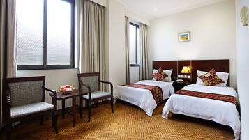 Quarto de hotel com duas camas, uma cadeira, uma mesinha com frutas e amplas janelas com cortinas.