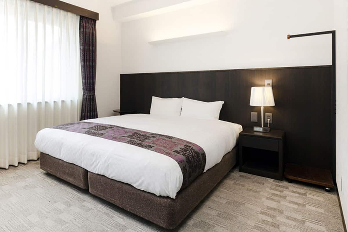 Una habitación de hotel con una cama grande, una mesita de noche con lámpara y una ventana con cortinas traslúcidas.