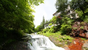 Une cascade d’eau se déversant sur des rochers, entourée d’une végétation luxuriante, et un bâtiment doté de balcons en arrière-plan.