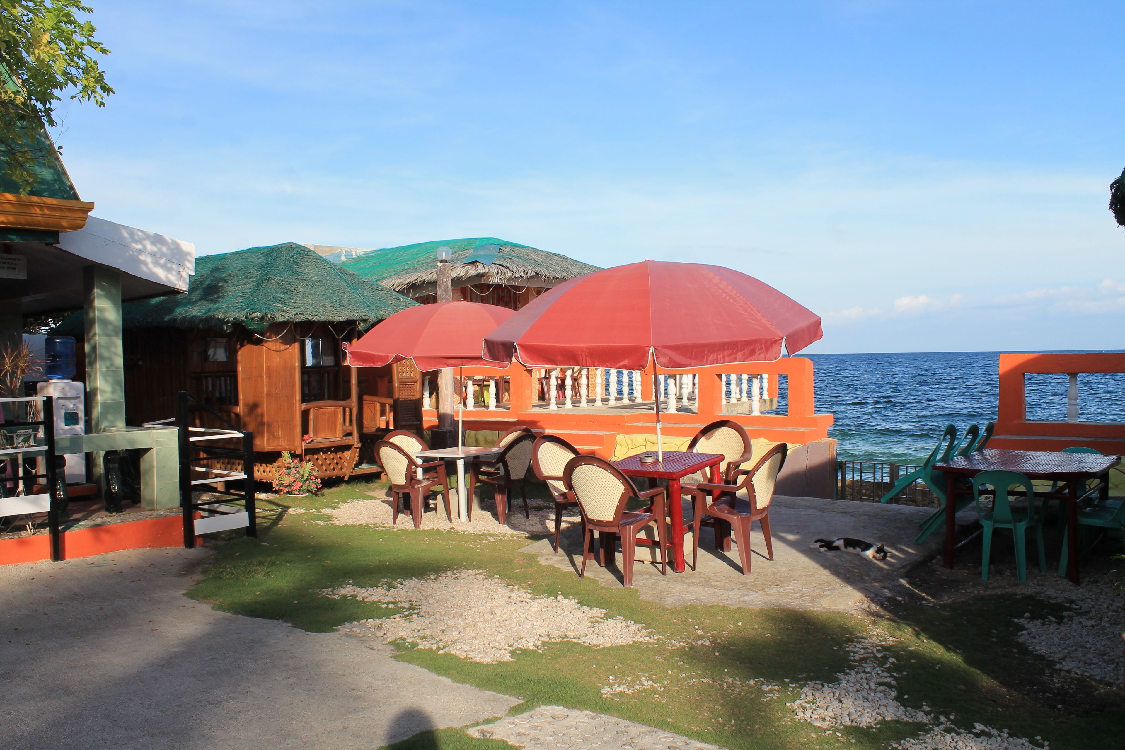 An outdoor seating area with tables and chairs, a thatched-roof structure, and a view of the sea.