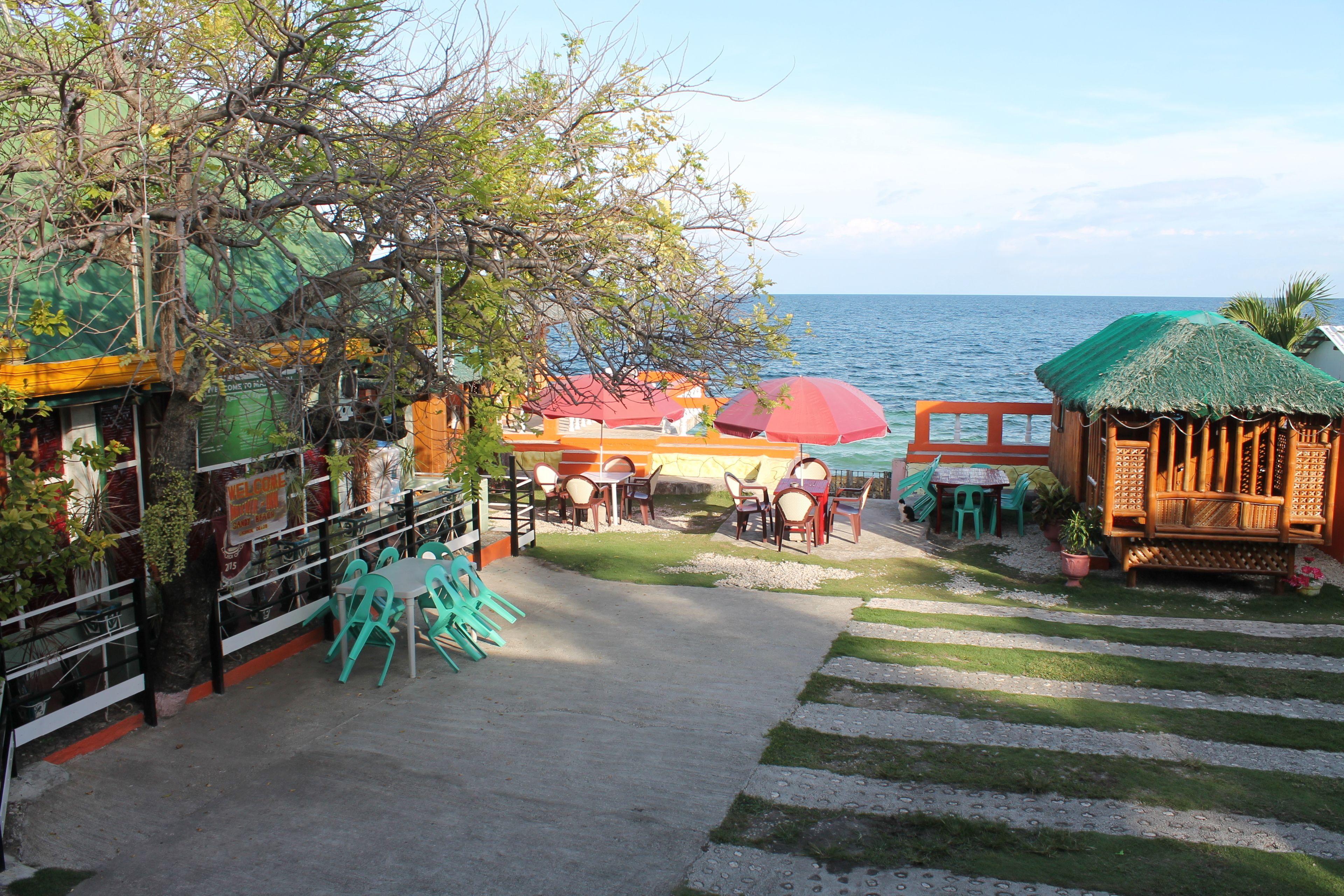A coastal restaurant with outdoor seating, a view of the sea, and a thatched-roof structure.