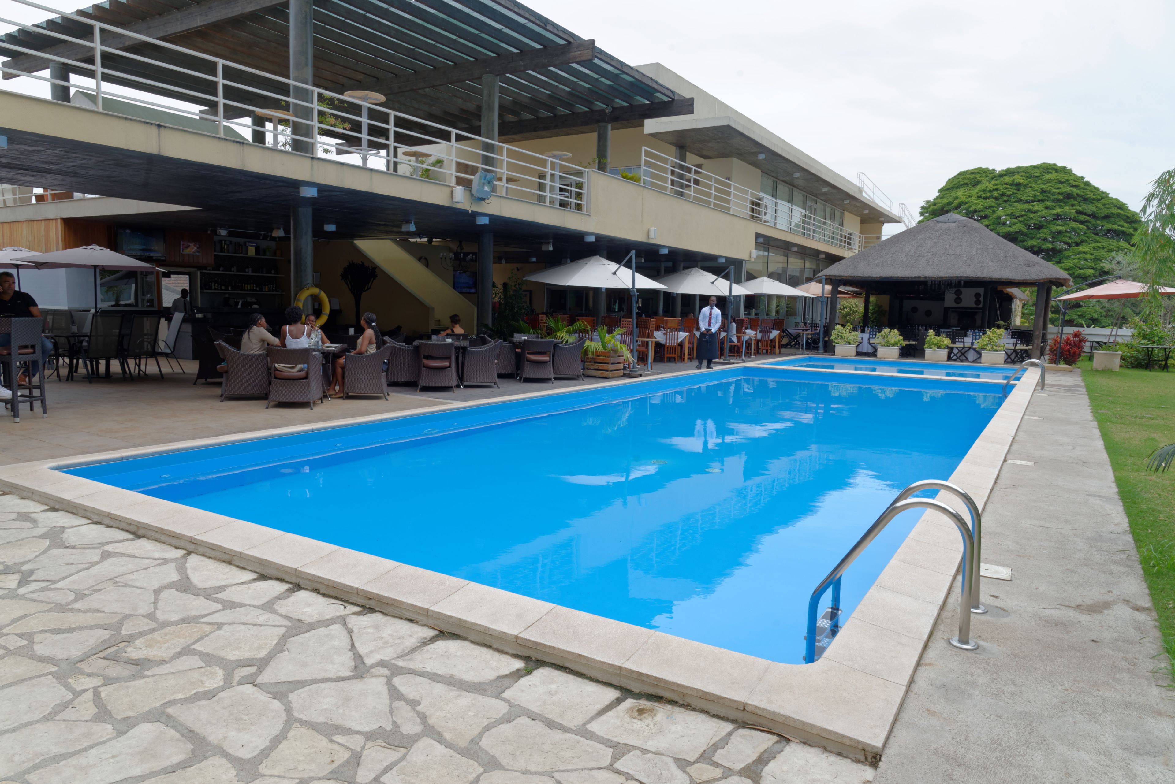 Uma piscina ao ar livre com piso de pedra, cercada por assentos e guarda-sóis, e um edifício com área de varanda coberta ao fundo.