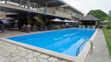 Uma piscina ao ar livre com piso de pedra, cercada por assentos e guarda-sóis, e um edifício com área de varanda coberta ao fundo.