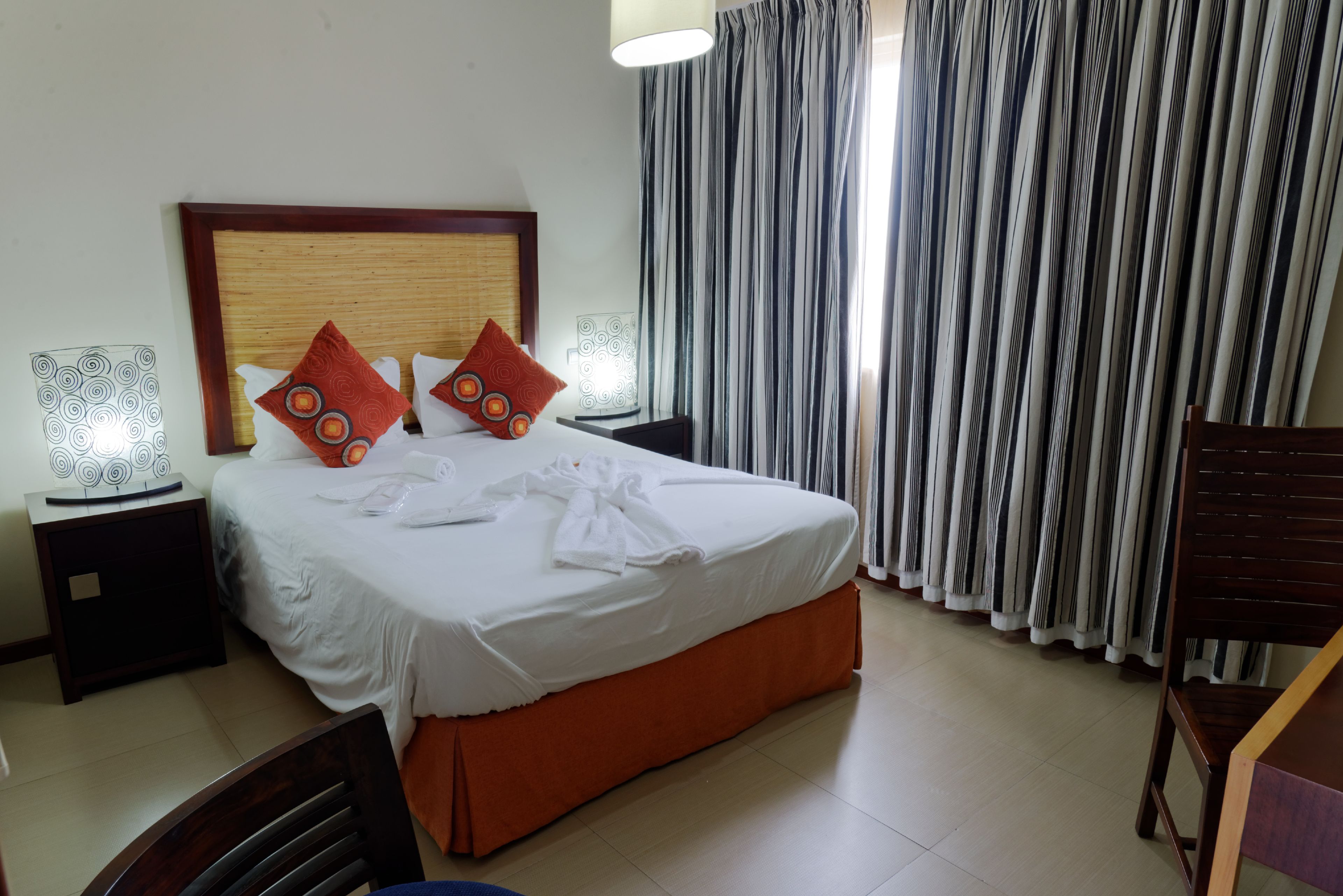 A neatly made bed with a wooden headboard, two bedside tables with lamps, a chair, and a large window with striped curtains.