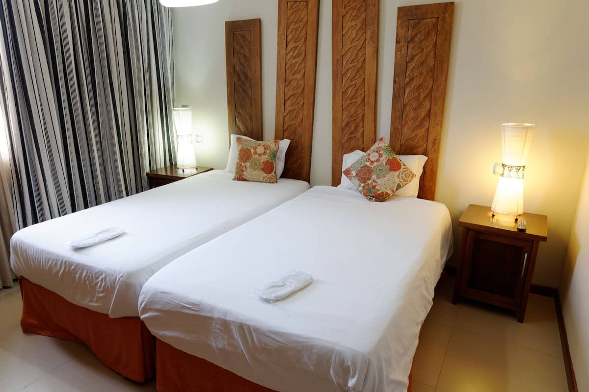 Ein Hotelzimmer mit zwei Betten, jeweils mit weißer Bettwäsche und dekorativen Kissen. Es gibt ein Holz-Kopfteil mit gewebtem Muster, einen Nachttisch mit Lampe sowie ein Fenster mit Vorhängen.
