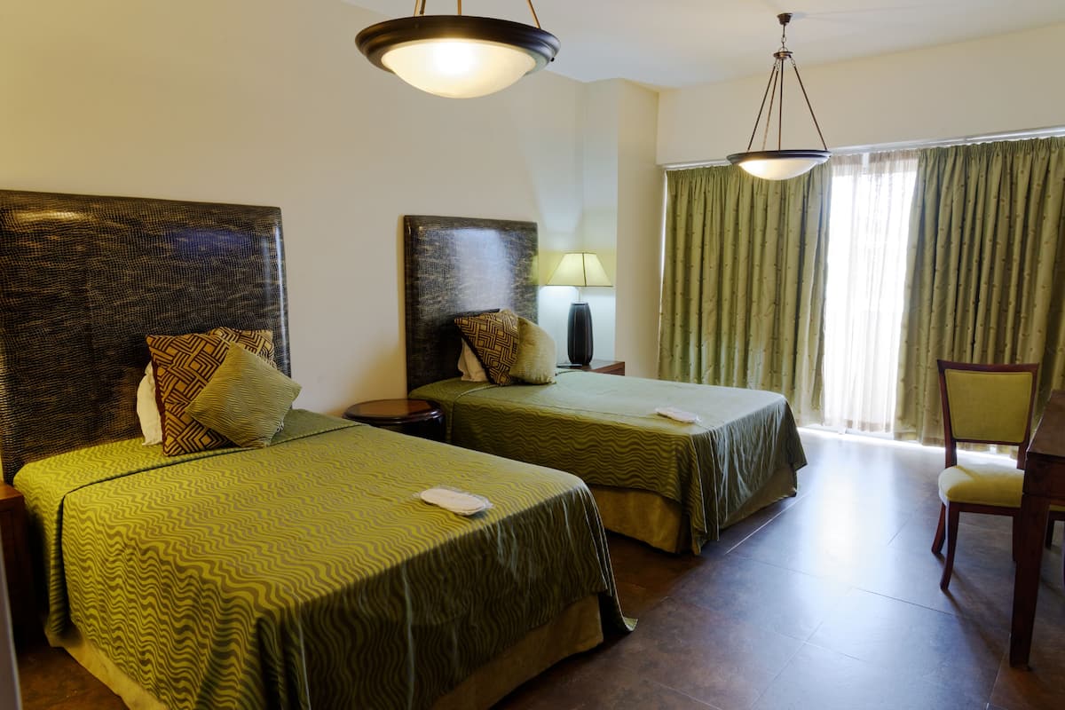 Ein Hotelzimmer mit zwei Betten, einem Sessel, einer Lampe und einem großen Fenster mit Vorhängen.
