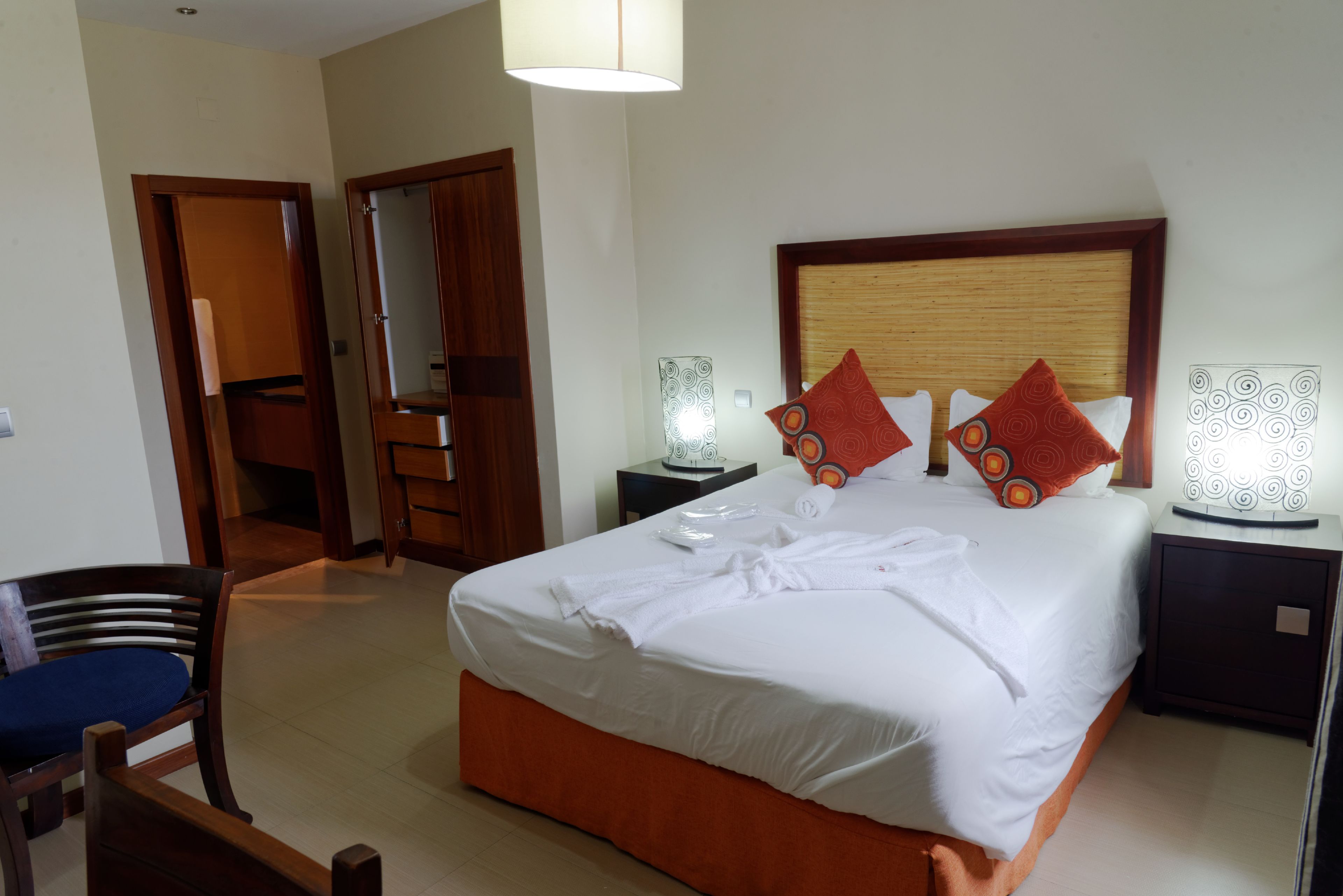 A hotel room with a large bed, a wooden headboard, two bedside tables with lamps, a chair, and a wardrobe.