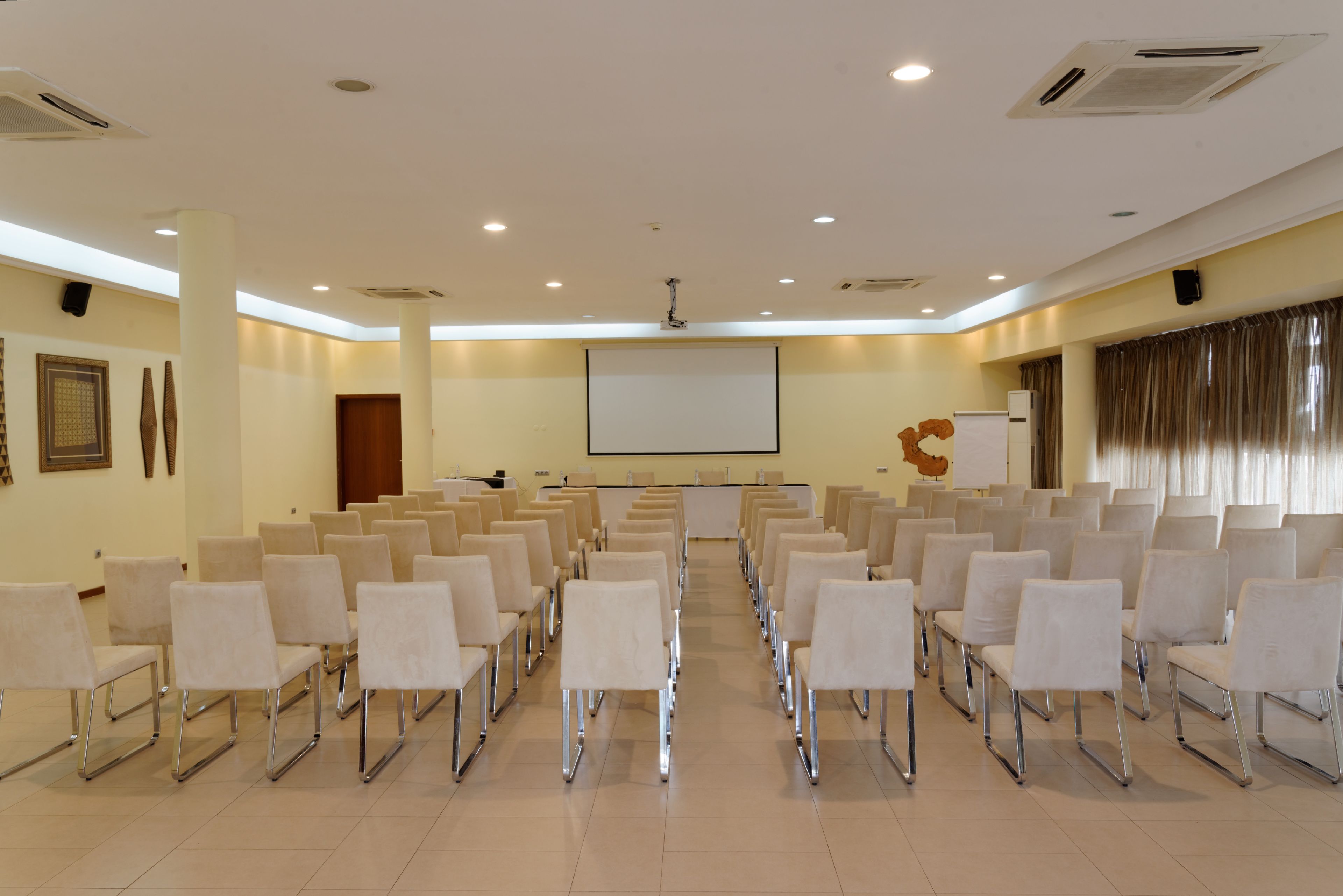 Un salón de conferencias con filas de sillas blancas, una pantalla de proyector y una pizarra.