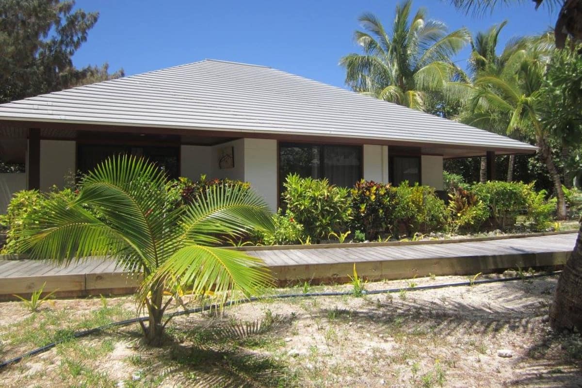 A two-story building with a flat roof, surrounded by tropical vegetation including palm trees and various shrubs.