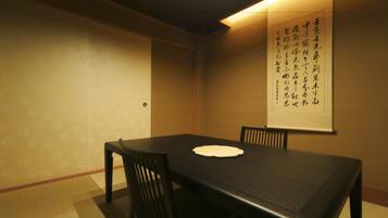 Una habitación japonesa tradicional con suelo de tatami, una mesa baja y dos sillas de madera. Un pergamino con caligrafía se exhibe en la pared.
