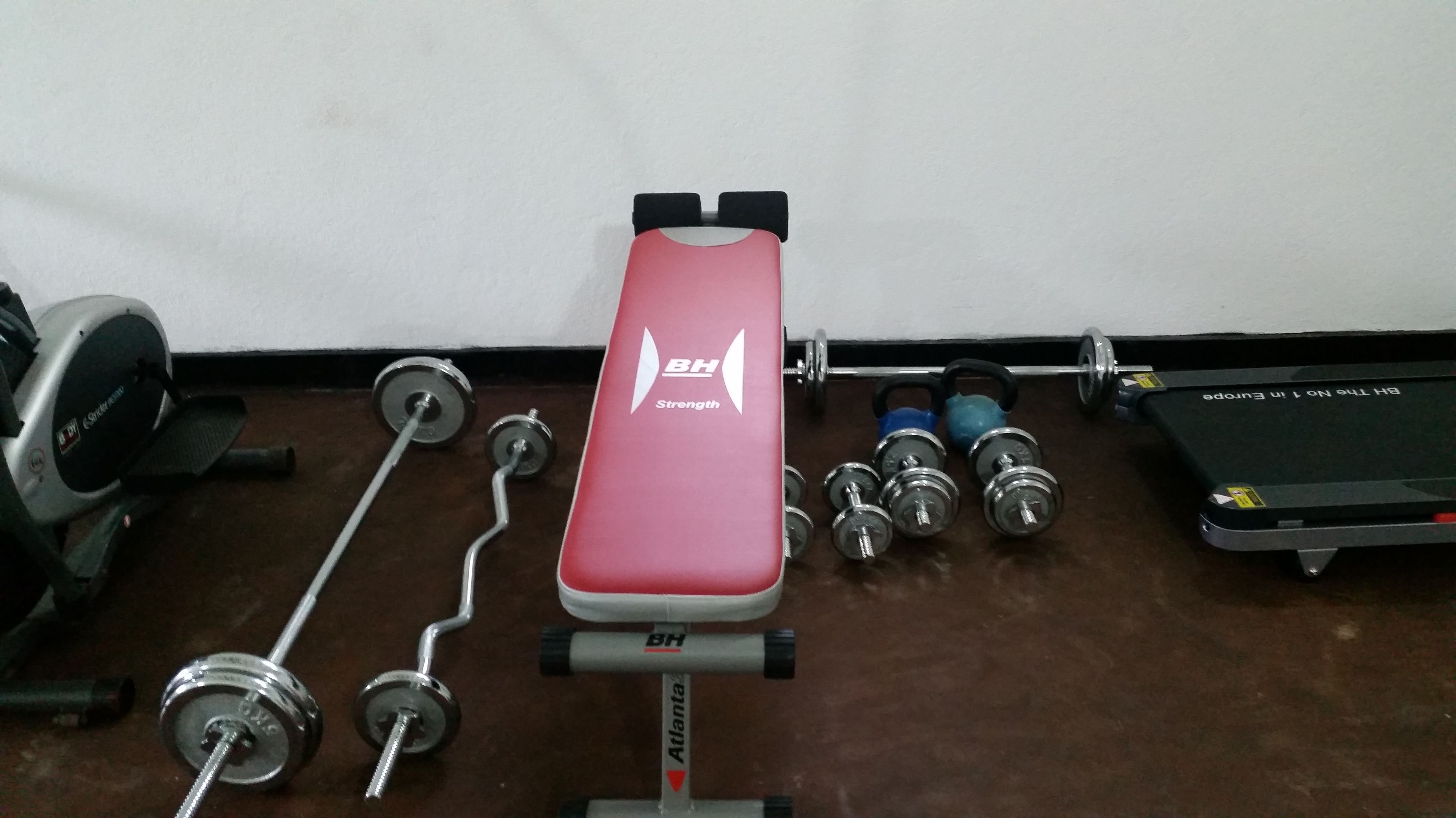 Un banco rojo con la marca 'BH Strength', una barra, mancuernas y un soporte para discos de pesas en un gimnasio.