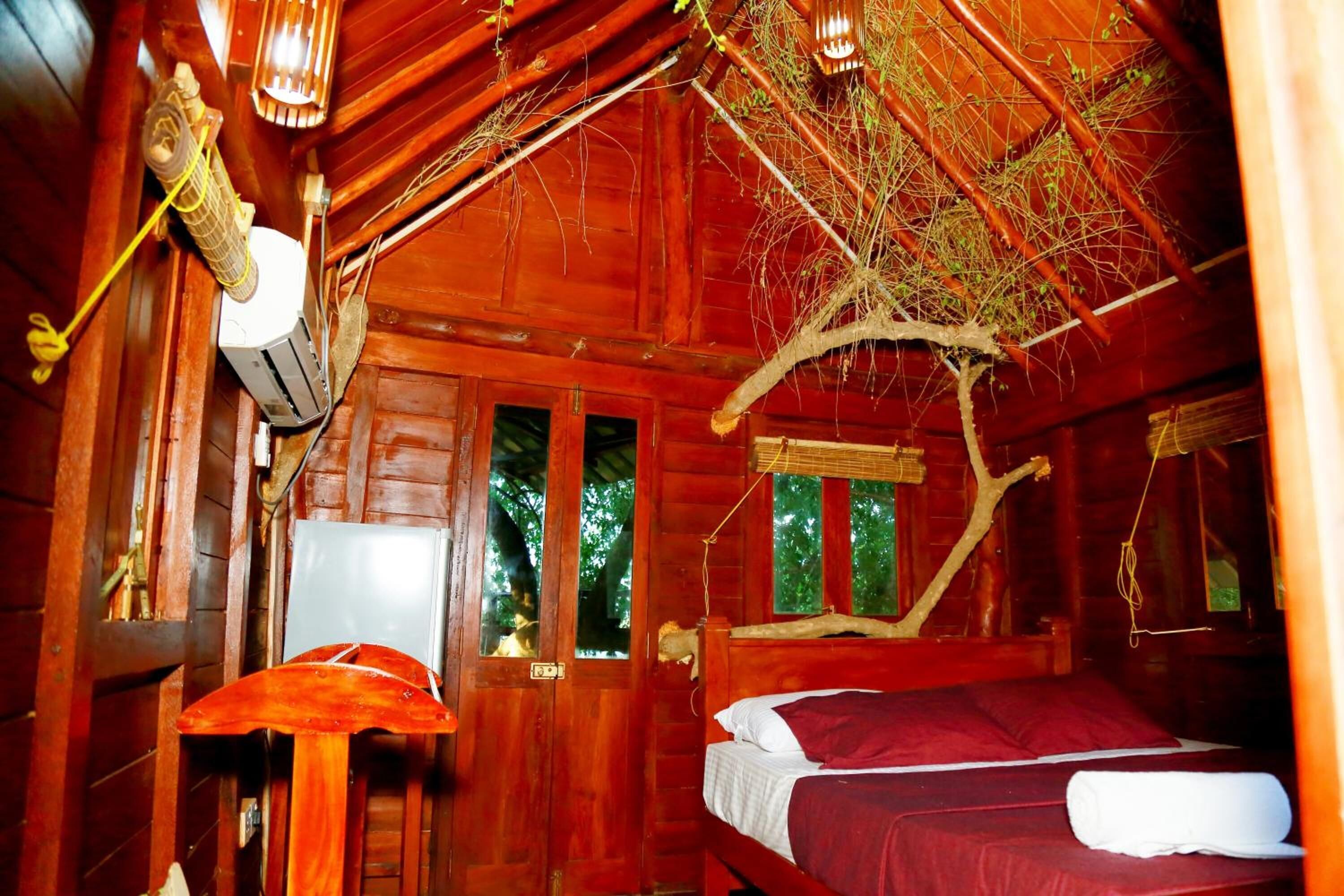 A wooden cabin room with a bed, a hanging light fixture, and a window with a view of trees.