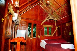 A wooden cabin room with a bed, a hanging light fixture, and a window with a view of trees.