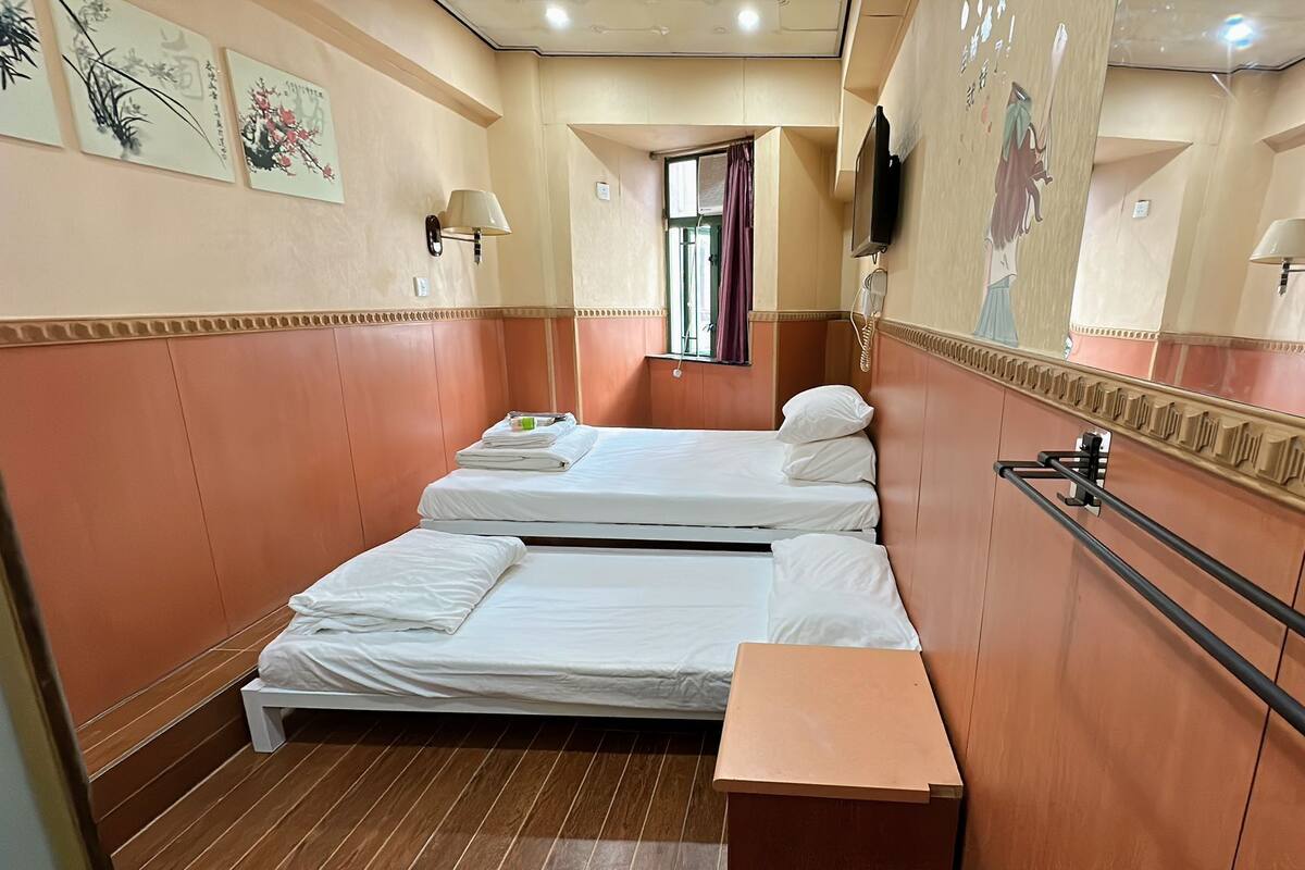 A hotel room with two beds, a small table, a wall-mounted TV, and artwork on the walls.