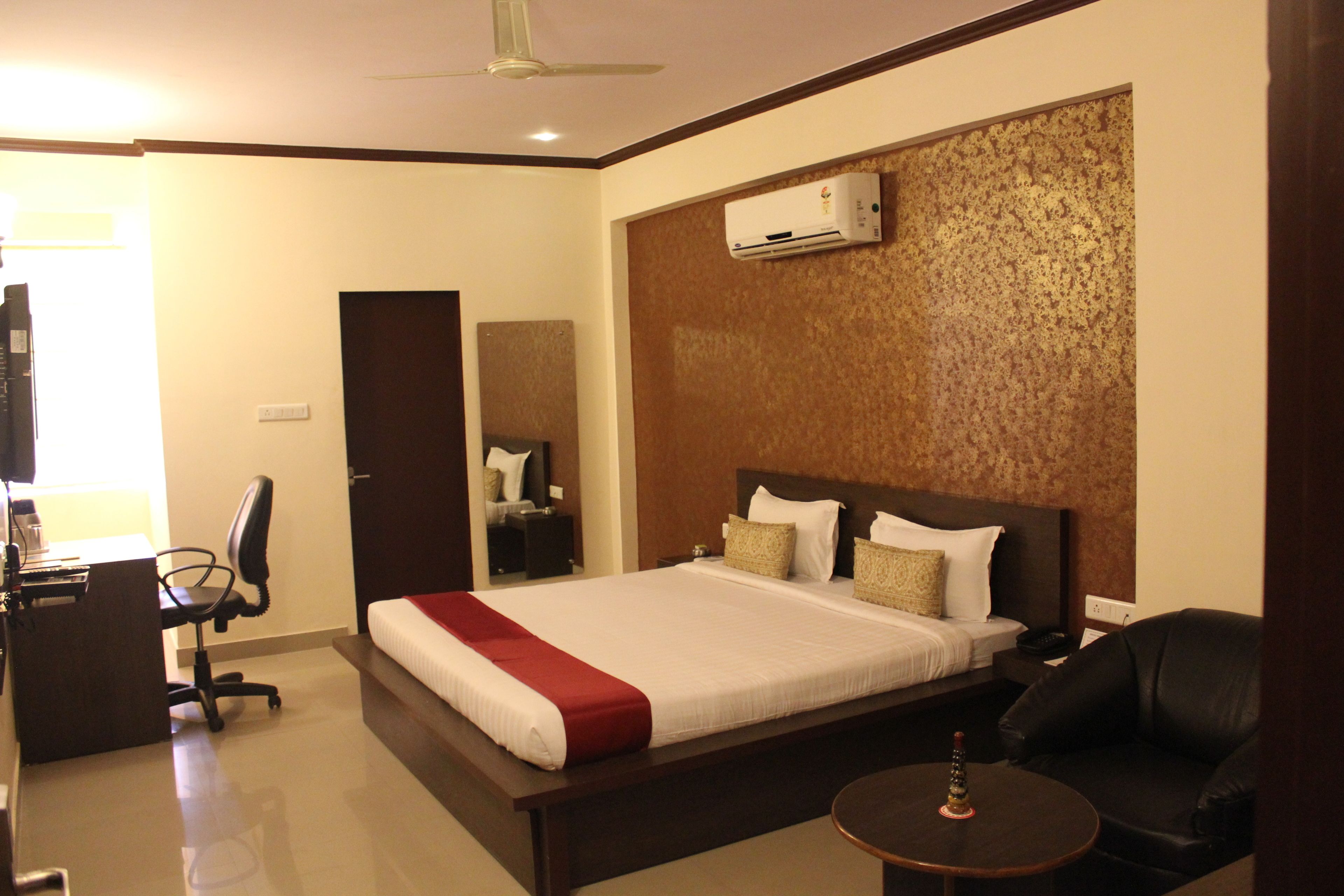 A hotel room with a large bed, a desk with a chair, a small round table, and a ceiling fan.