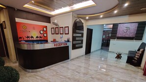 A hotel reception area with a reception desk labeled "RECEPTION," a clock, framed certificates, and a wall with a logo and text. - Hotel Savi Regency (Jaipur)