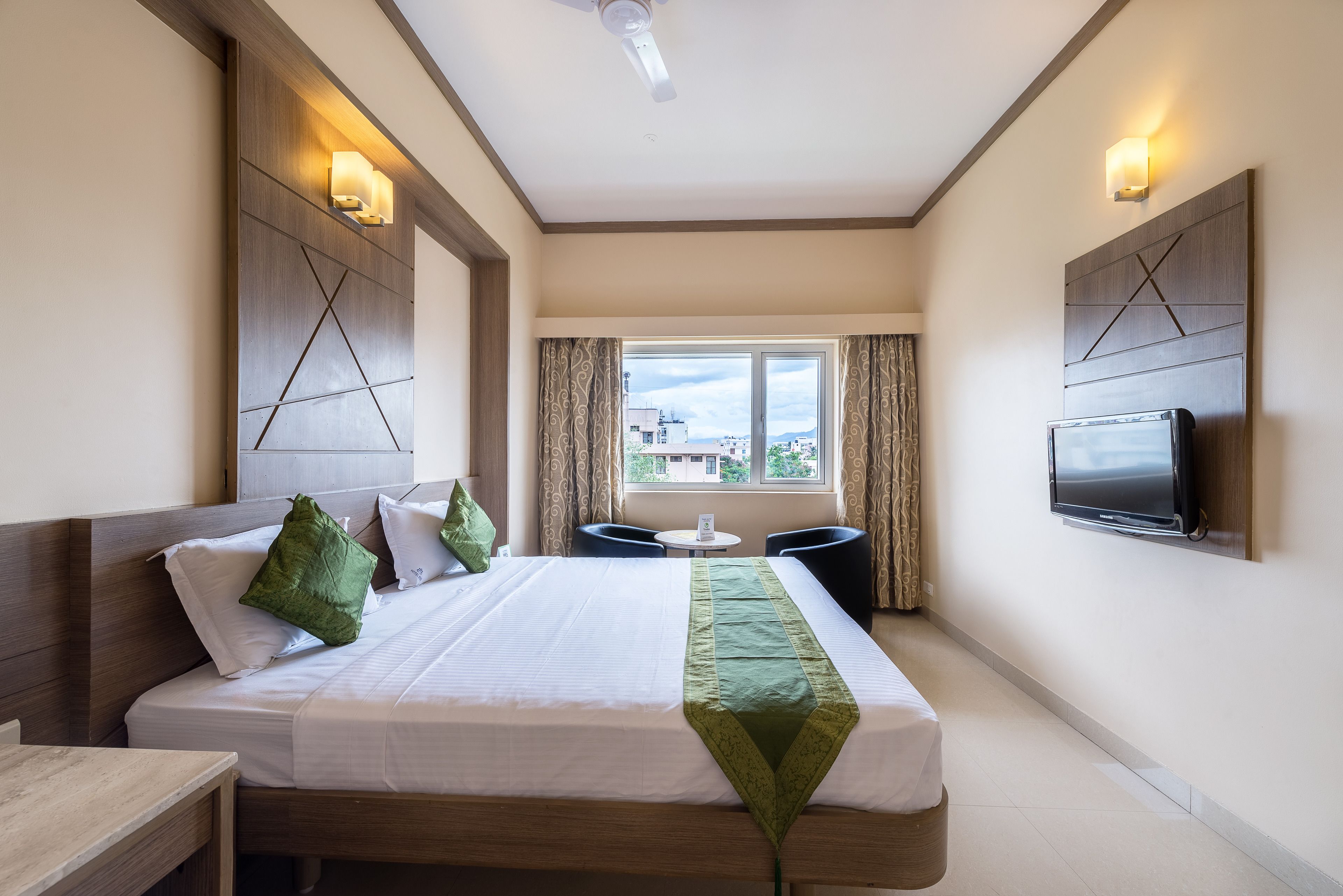 A hotel room with a large bed, two bedside tables, a small round table, a TV mounted on the wall, and a window with curtains.