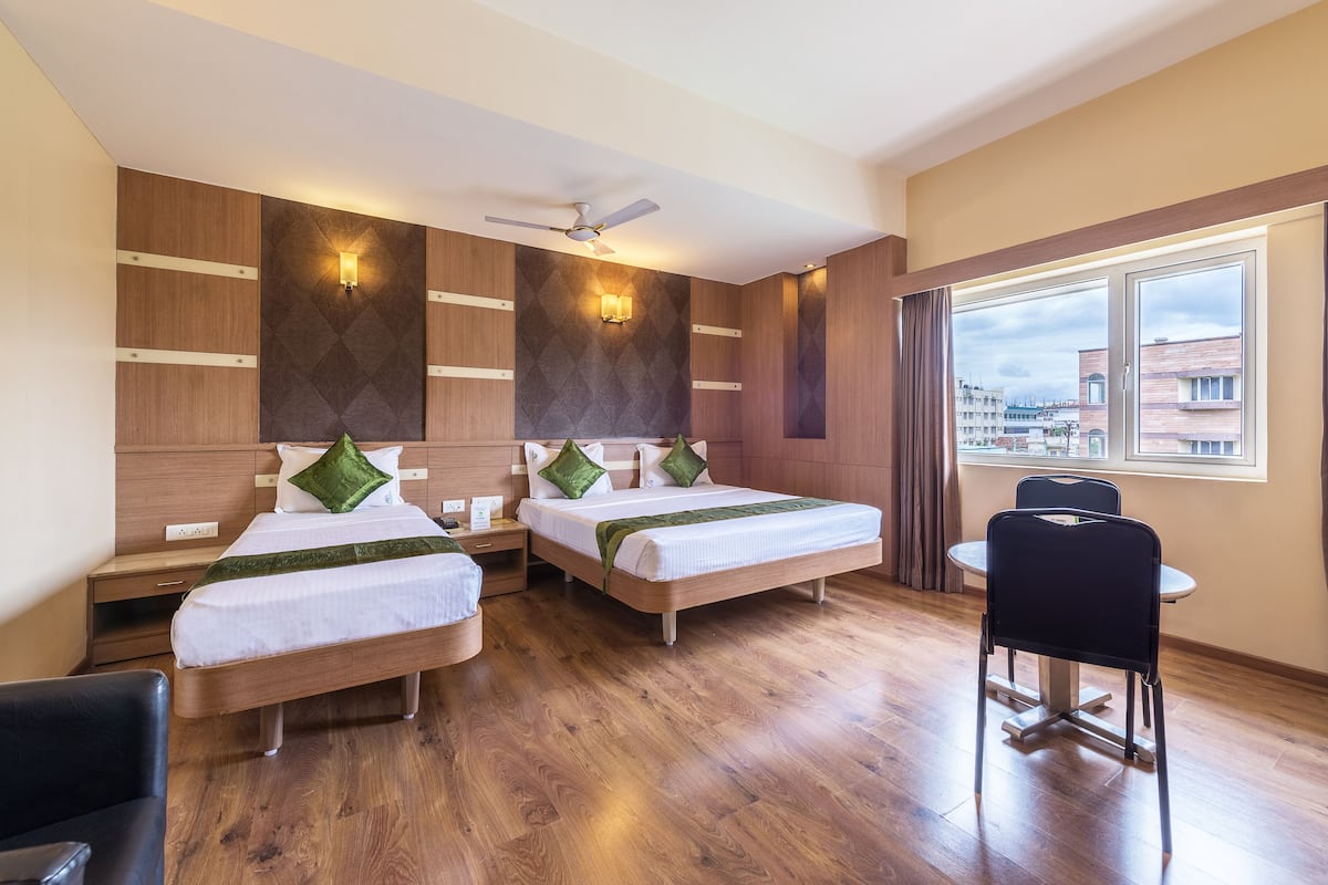 Habitación de hotel con dos camas, un ventilador de techo, una mesa de comedor con una silla y una ventana con vistas a edificios.