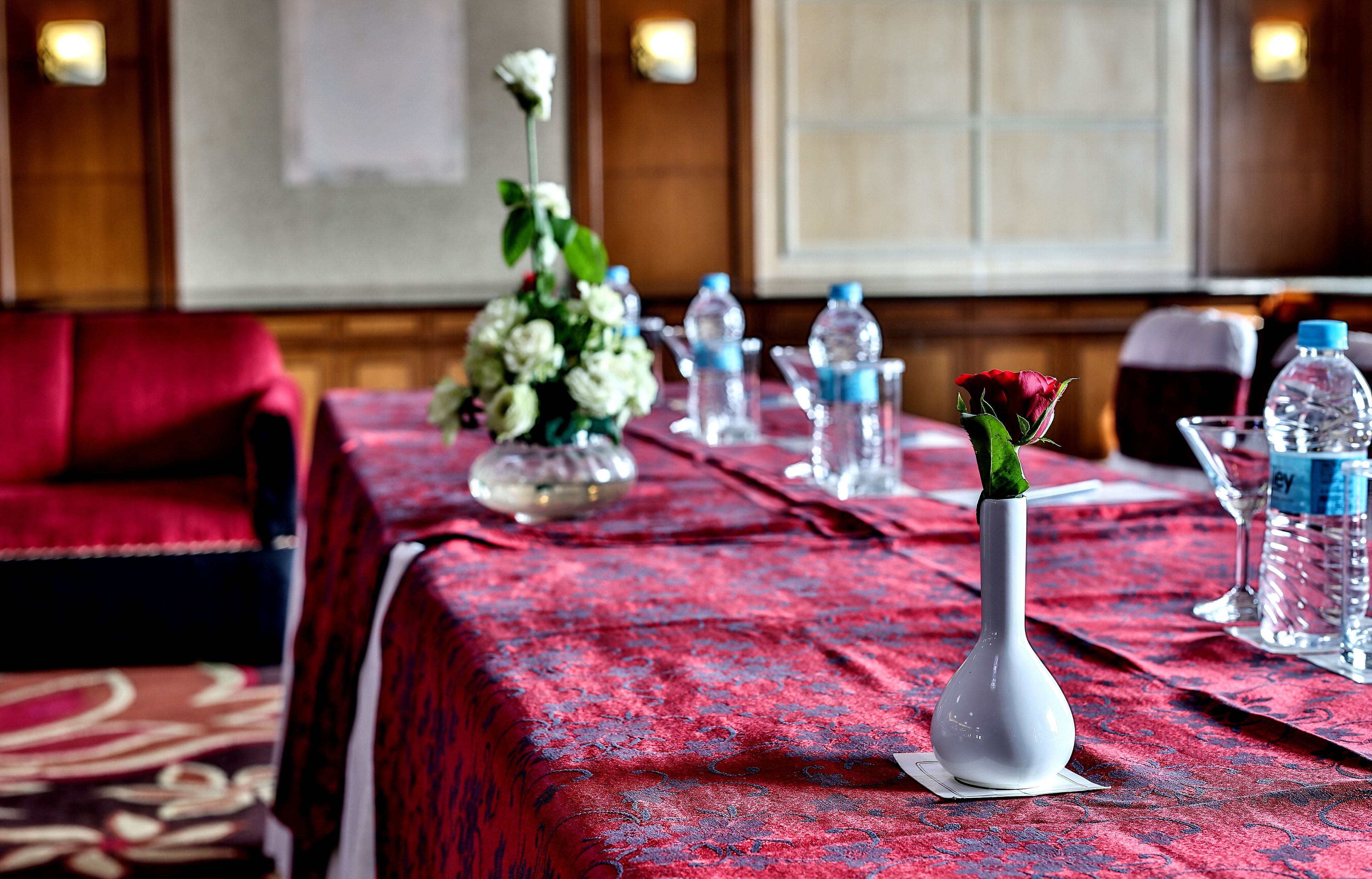 A conference table with a red patterned tablecloth, water bottles, a white vase with a rose, and a floral arrangement.