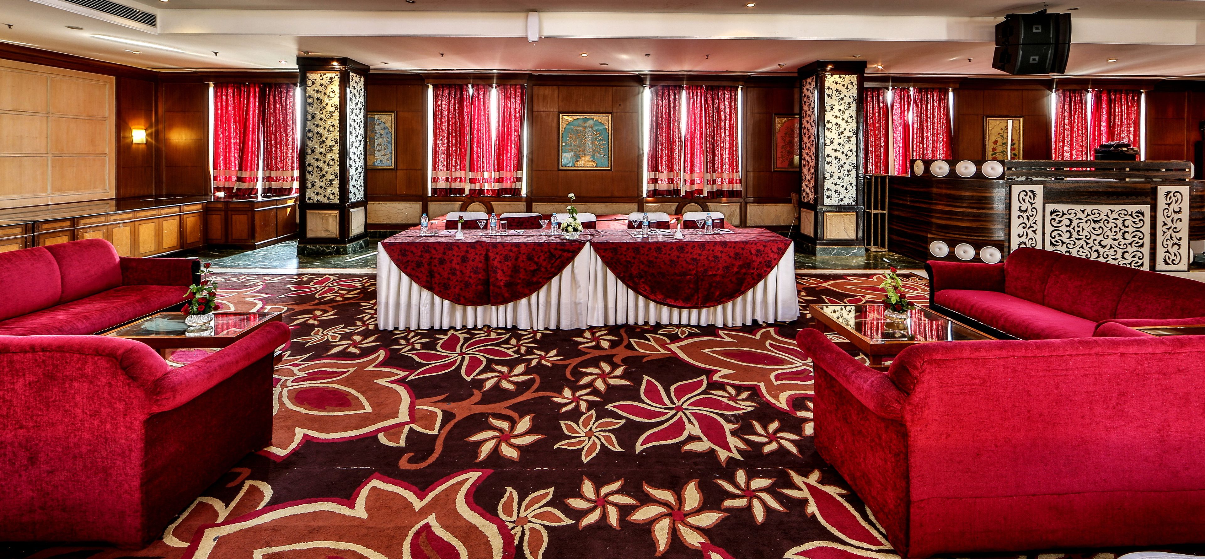 Un vestíbulo de hotel con alfombra roja, dos mesas redondas con manteles blancos, sofás rojos y un mostrador de recepción.