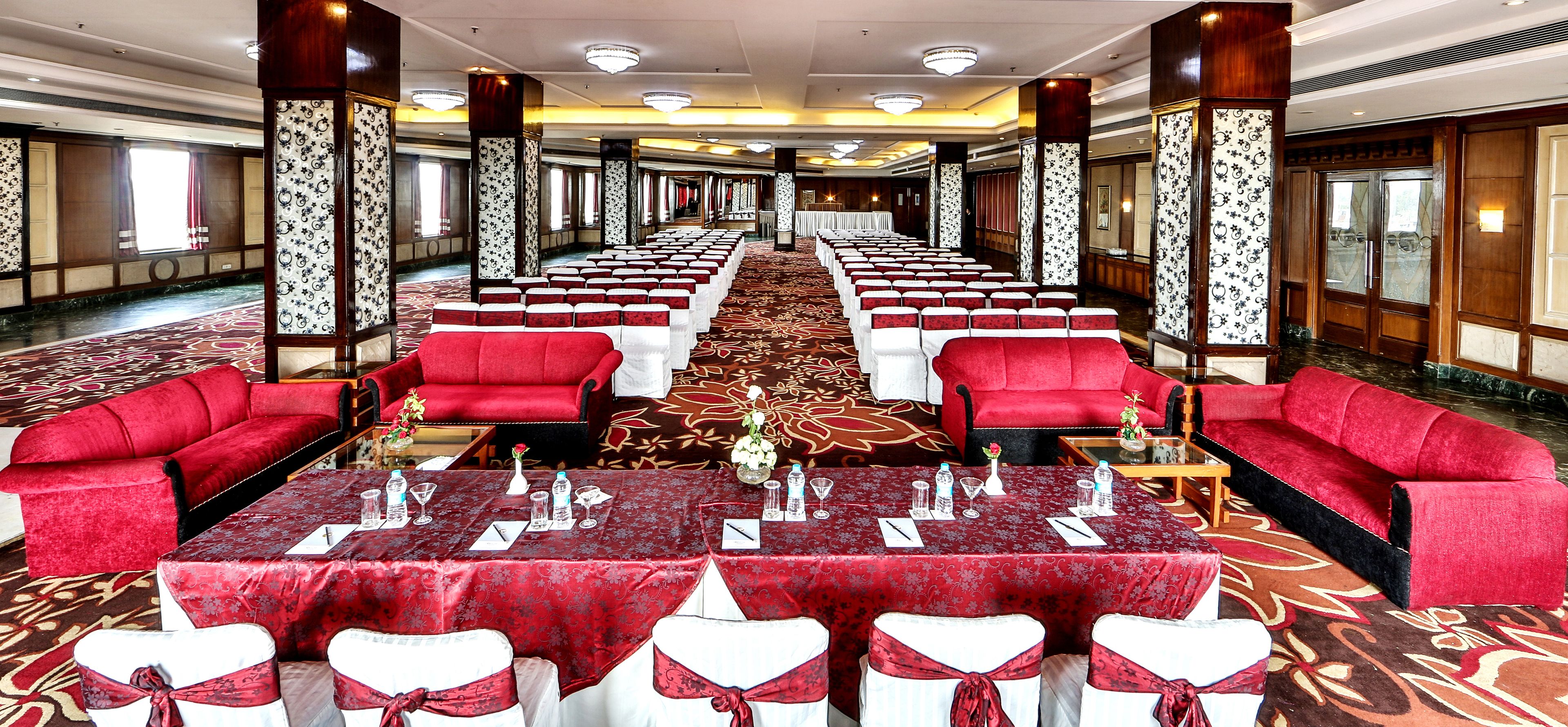 Un salón de banquetes con filas de sillas tapizadas en rojo, mesas blancas con manteles rojos y una alfombra estampada.