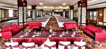 Un salón de banquetes con filas de sillas tapizadas en rojo, mesas blancas con manteles rojos y una alfombra estampada.