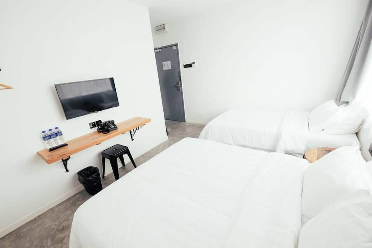 A hotel room with two beds, a TV on the wall, a wooden shelf with water bottles, and a door in the background.