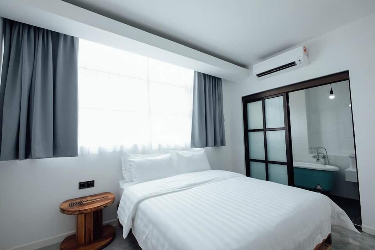 A modern hotel room with a large bed, a small wooden stool, a window with curtains, an air conditioning unit, and a bathroom with a bathtub and sink.