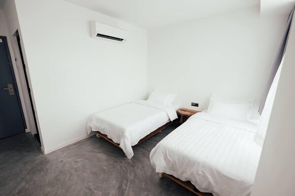 Una habitación pequeña y limpia con dos camas individuales, una mesita de noche de madera y un aire acondicionado instalado en la pared.