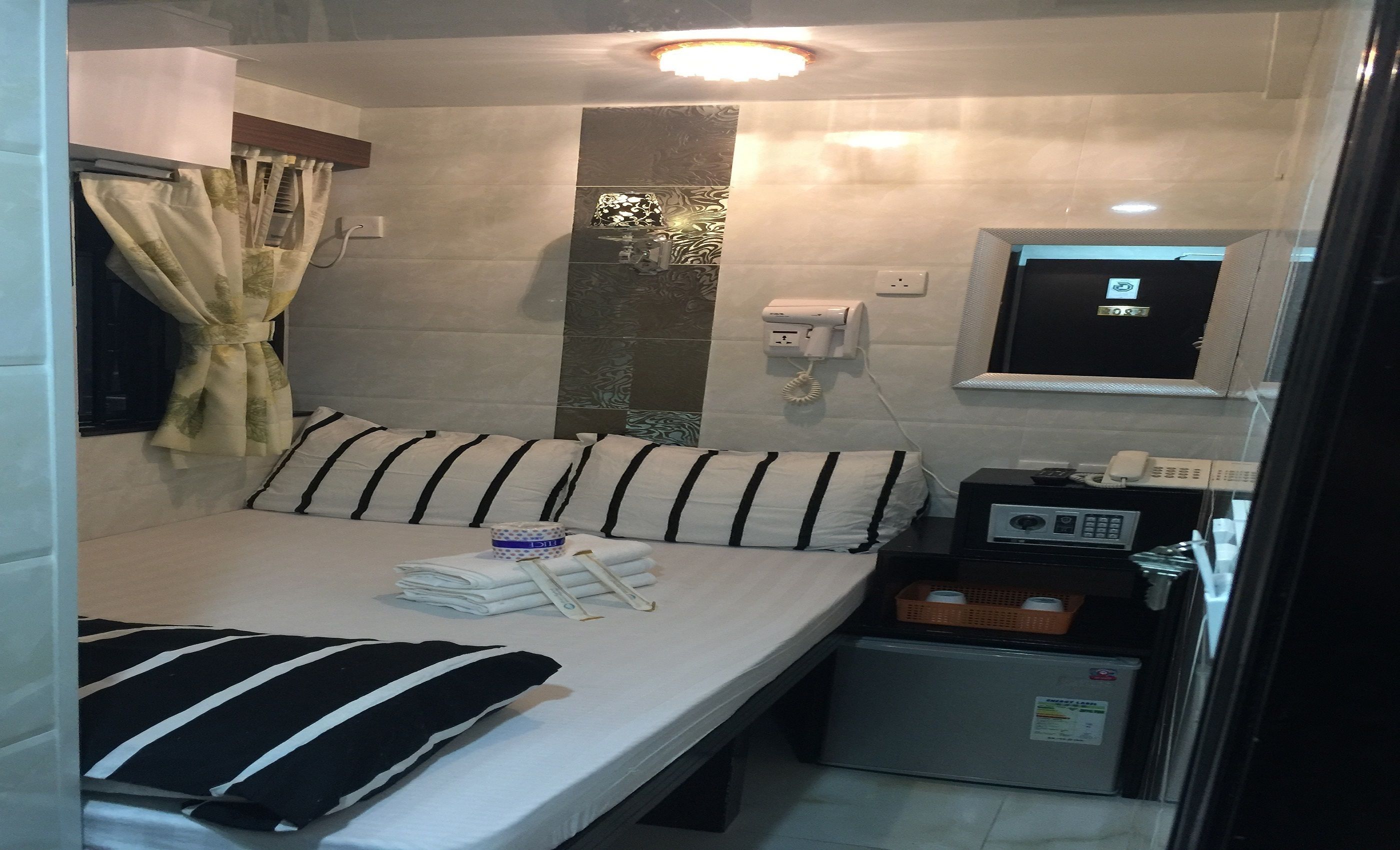 A compact hotel room with a bed, a small refrigerator, a bedside table with a radio, a telephone, and a wall-mounted TV.