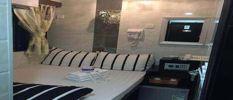 A compact hotel room with a bed, a small refrigerator, a bedside table with a radio, a telephone, and a wall-mounted TV.