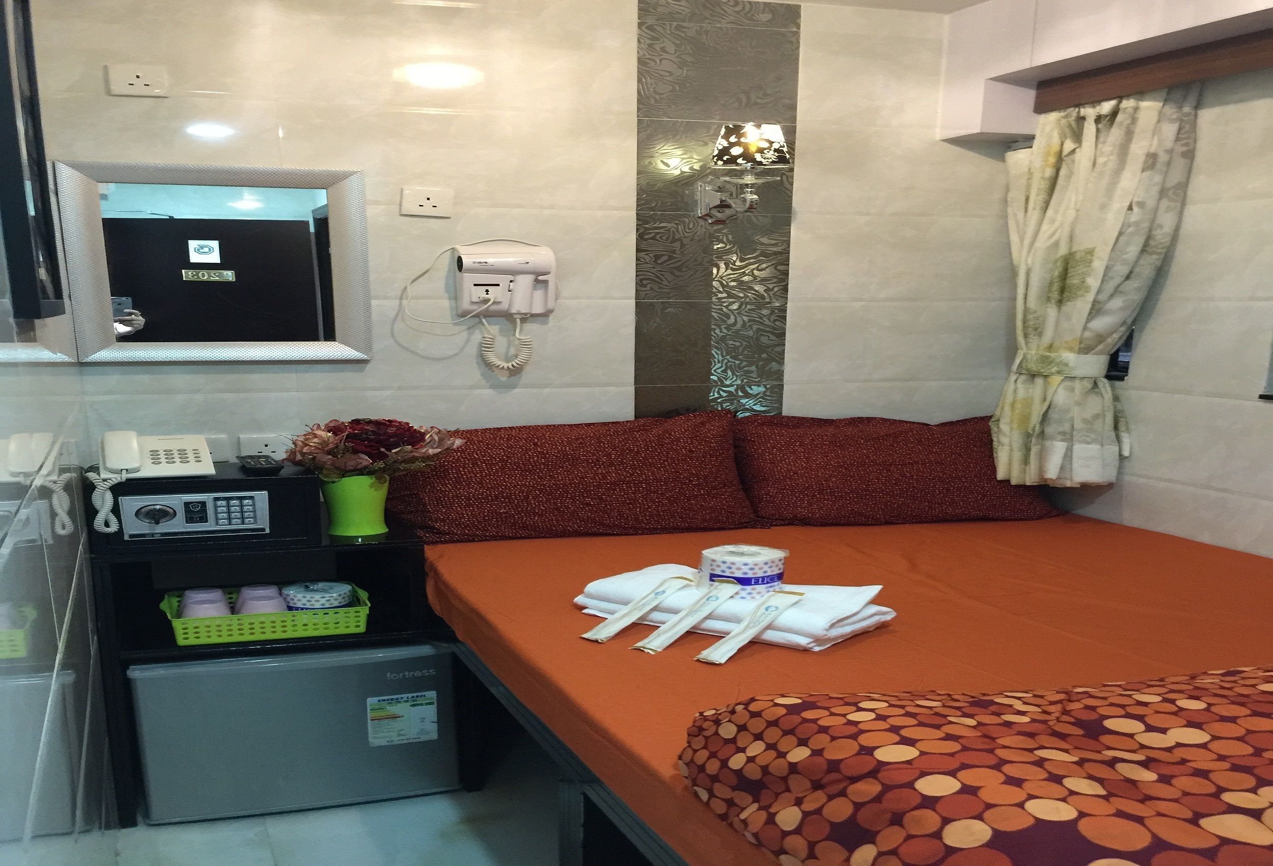 A hotel room with a bed, a small desk with a phone and a lamp, a mirror, and a mini-fridge.