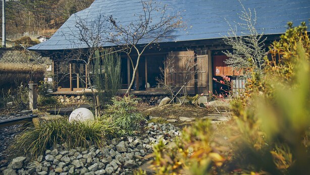 Ein traditionelles japanisches Haus mit geneigtem Dach, Holzfassade und einem Garten mit Steinen und wenig Bepflanzung.