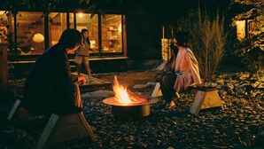 Ein Sitzbereich im Freien mit Feuerstelle, umgeben von Steinen und Pflanzen, mit einem Gebäude im Hintergrund.