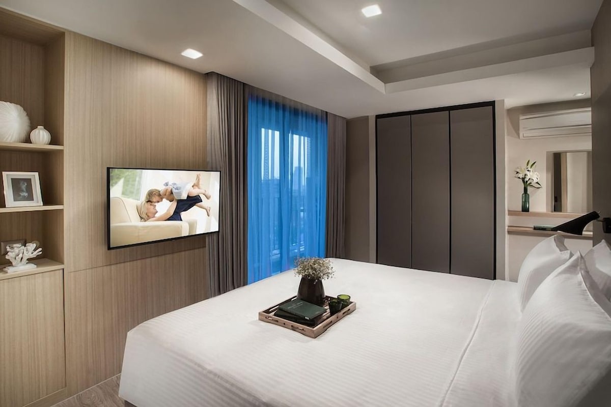 Ein modernes Hotelzimmer mit einem großen Bett, einem flachbildfernseher an der Wand, einem Nachttisch mit einer Vase und einem Kleiderschrank.