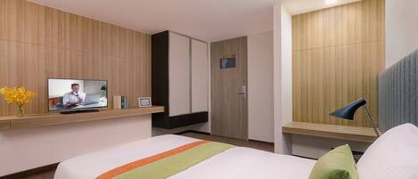 Habitación de hotel con cama, televisión sobre una estantería, puerta y escritorio con lámpara.