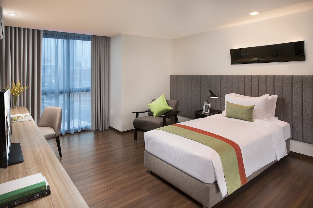 Una habitación de hotel moderna con una cama grande, un escritorio con silla, una mesita de noche con lámpara y un ventanal con cortinas.