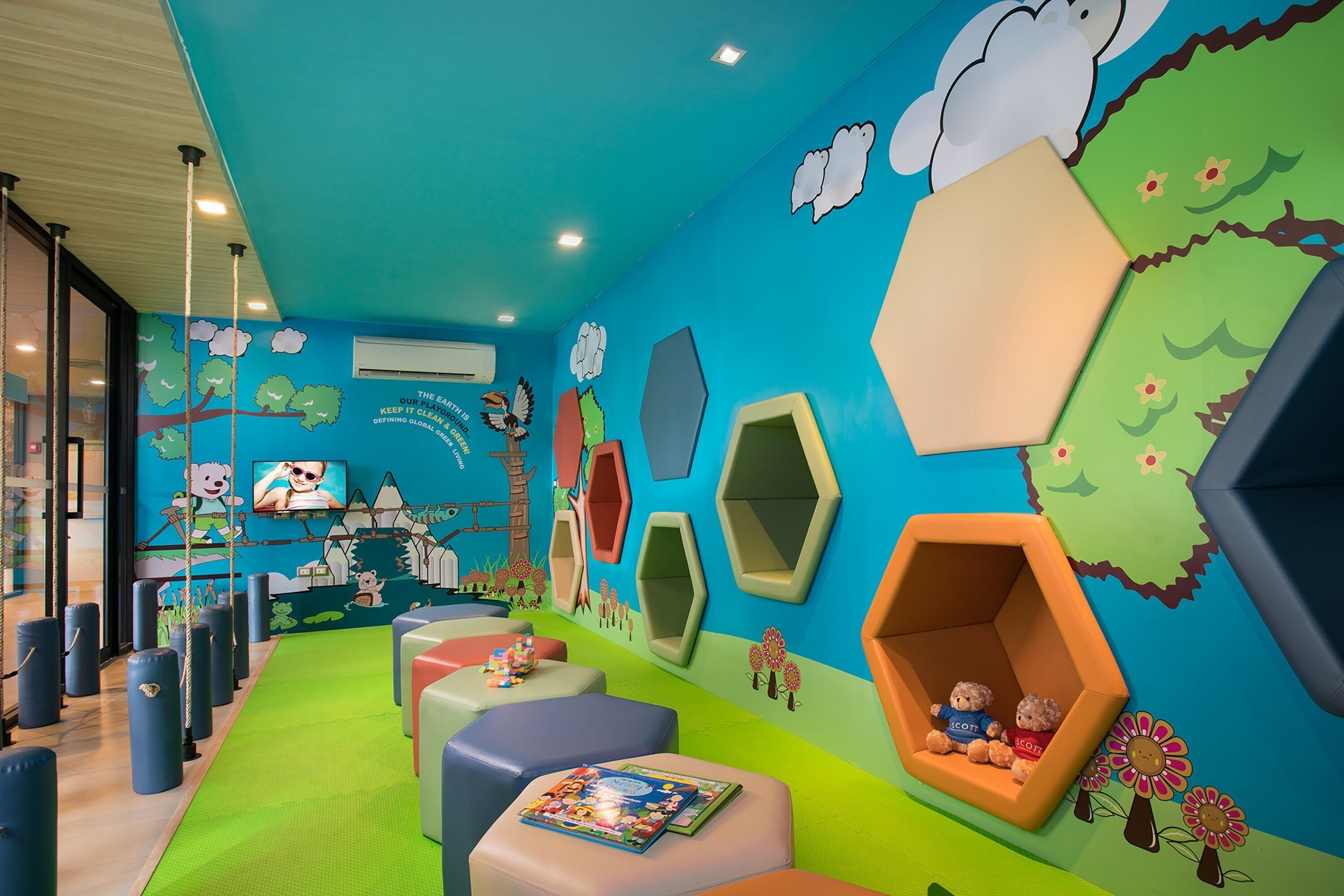 Zona de juegos infantiles con asientos hexagonales de colores, alfombra verde y decoraciones lúdicas en las paredes, incluyendo nubes y flores.