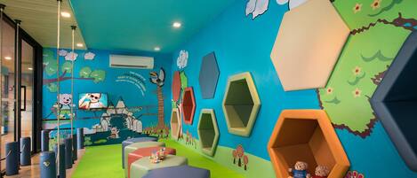 Zona de juegos infantiles con asientos hexagonales de colores, alfombra verde y decoraciones lúdicas en las paredes, incluyendo nubes y flores.