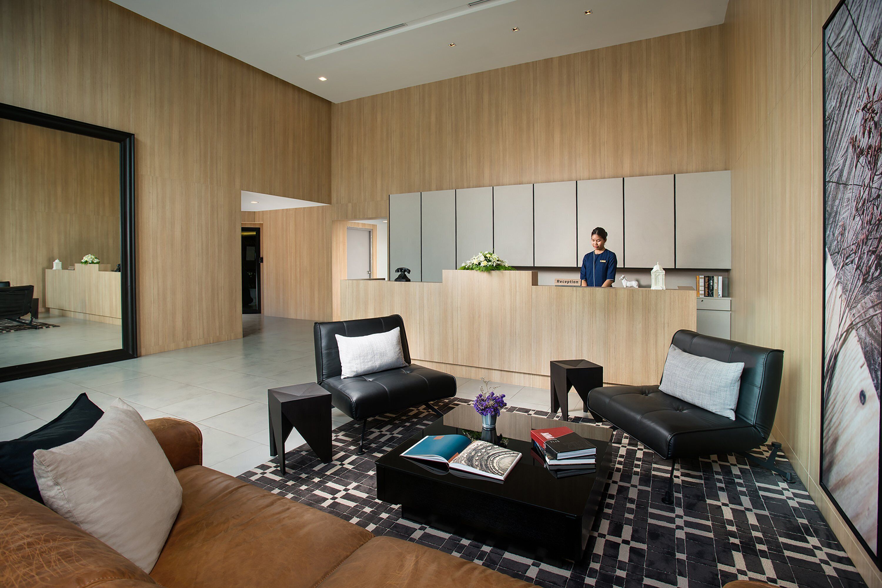 A modern hotel lobby with a reception desk, leather sofas, a coffee table with books, and a large mirror.