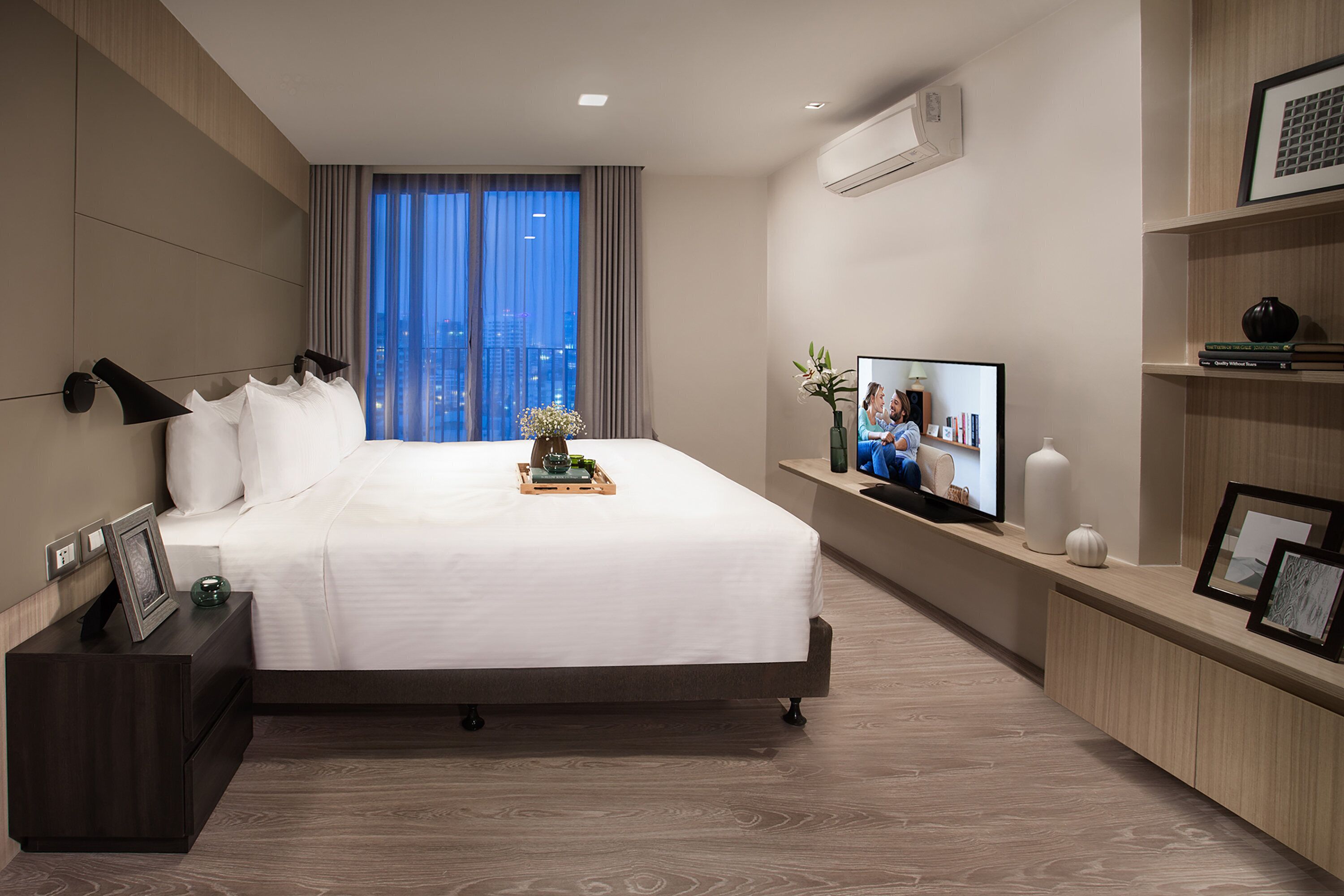 Ein modernes Hotelzimmer mit einem großen Bett, einem Fernseher auf einem Standfuß, einem Nachttisch und Blick auf die Stadt durch das Fenster.