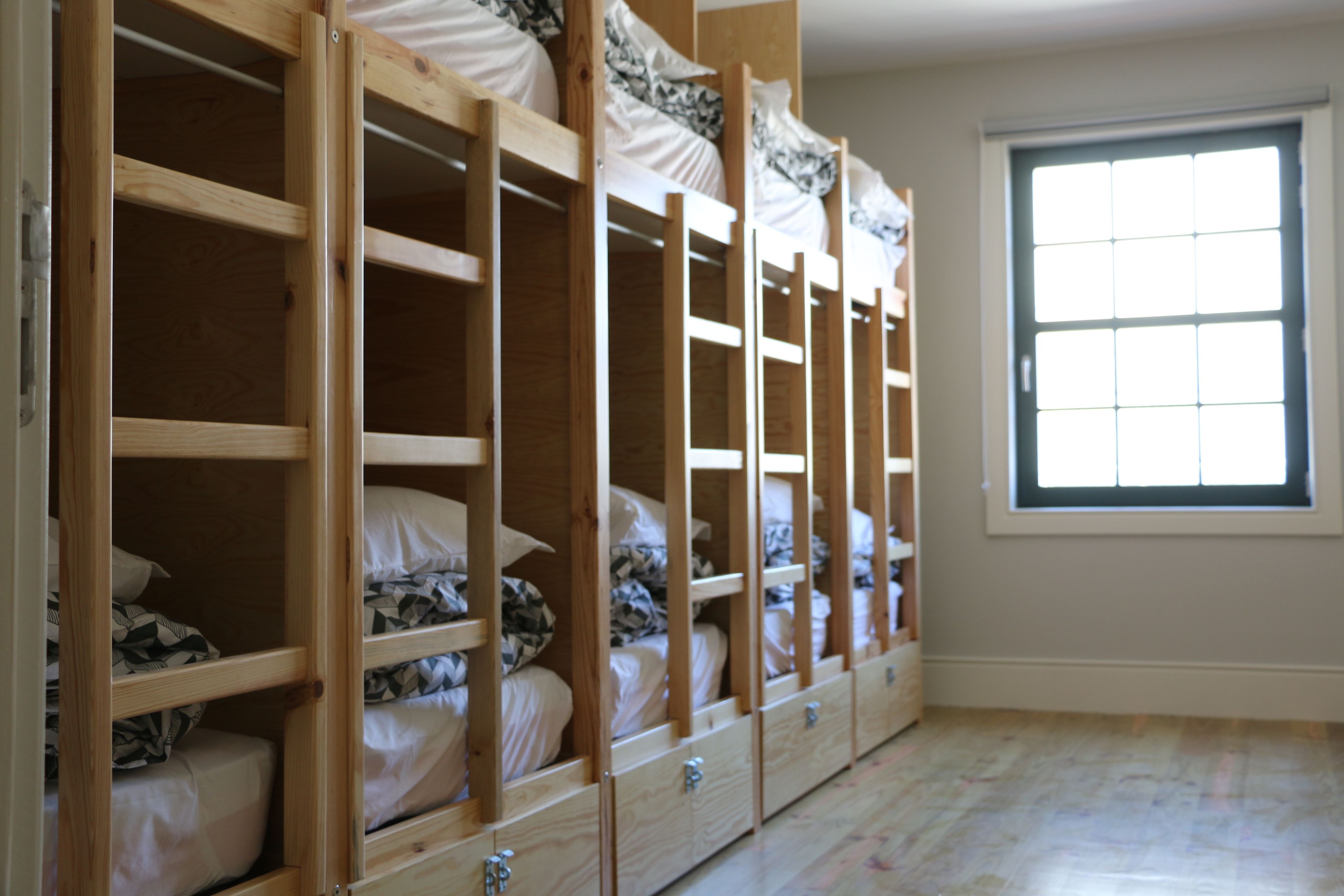 A room with bunk beds arranged in a row, a window with blinds, and a wooden floor.