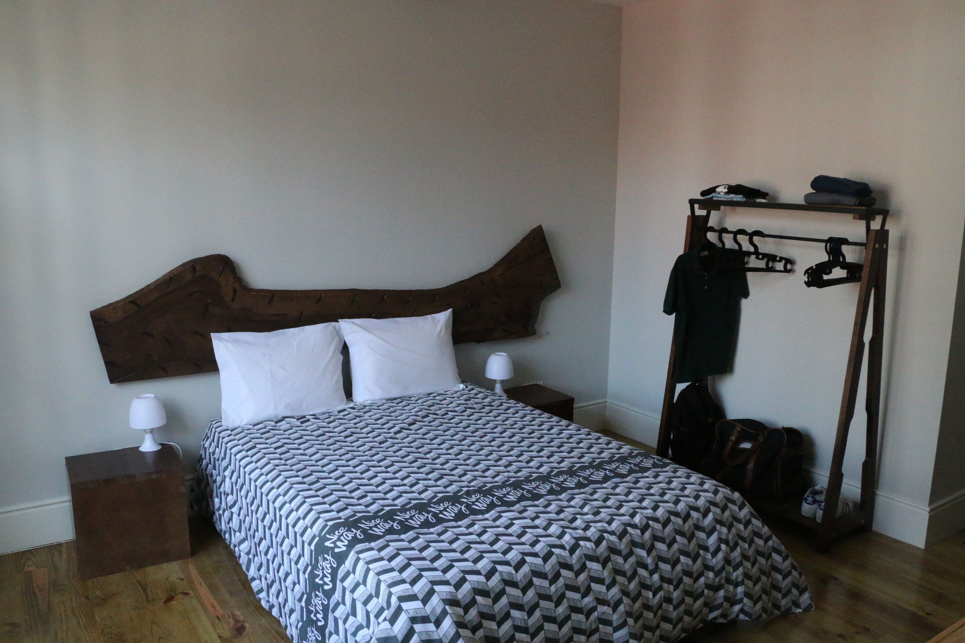 A neatly made bed with a patterned bedspread, a wooden headboard, a bedside table with a lamp, and a clothing rack against the wall.