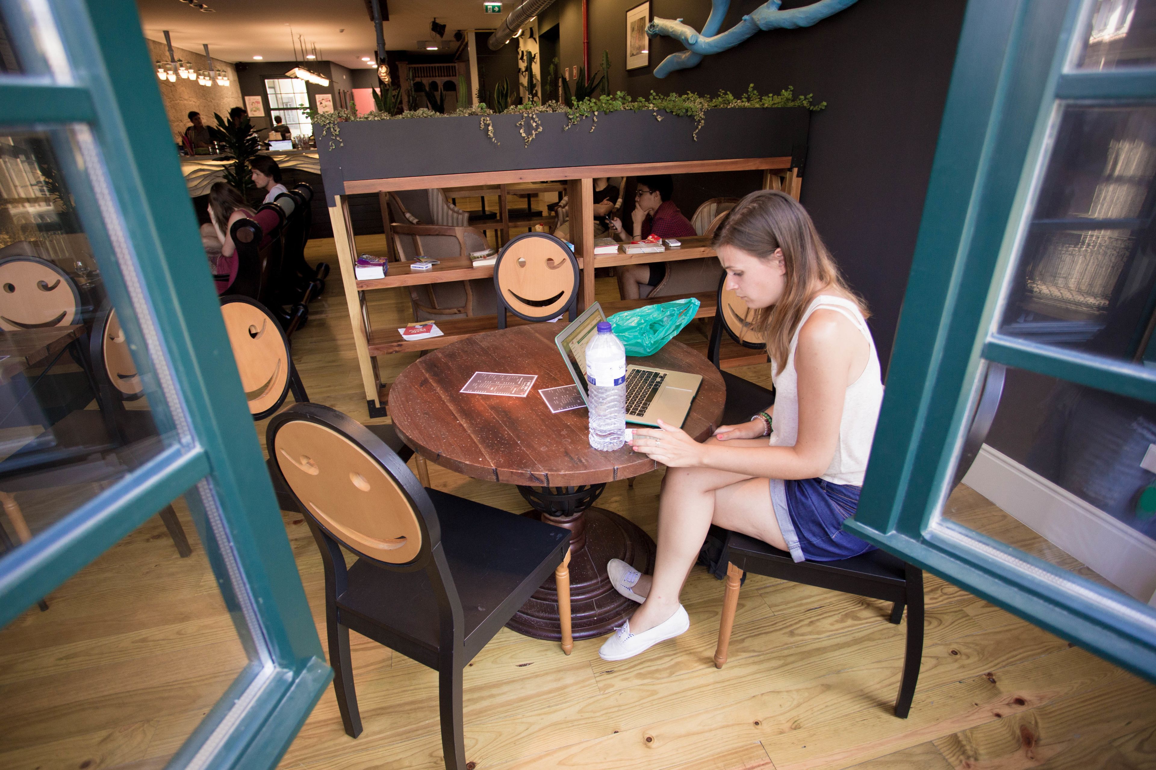A woman sitting at a round wooden table with a laptop, surrounded by wooden chairs and a smiley face chair.