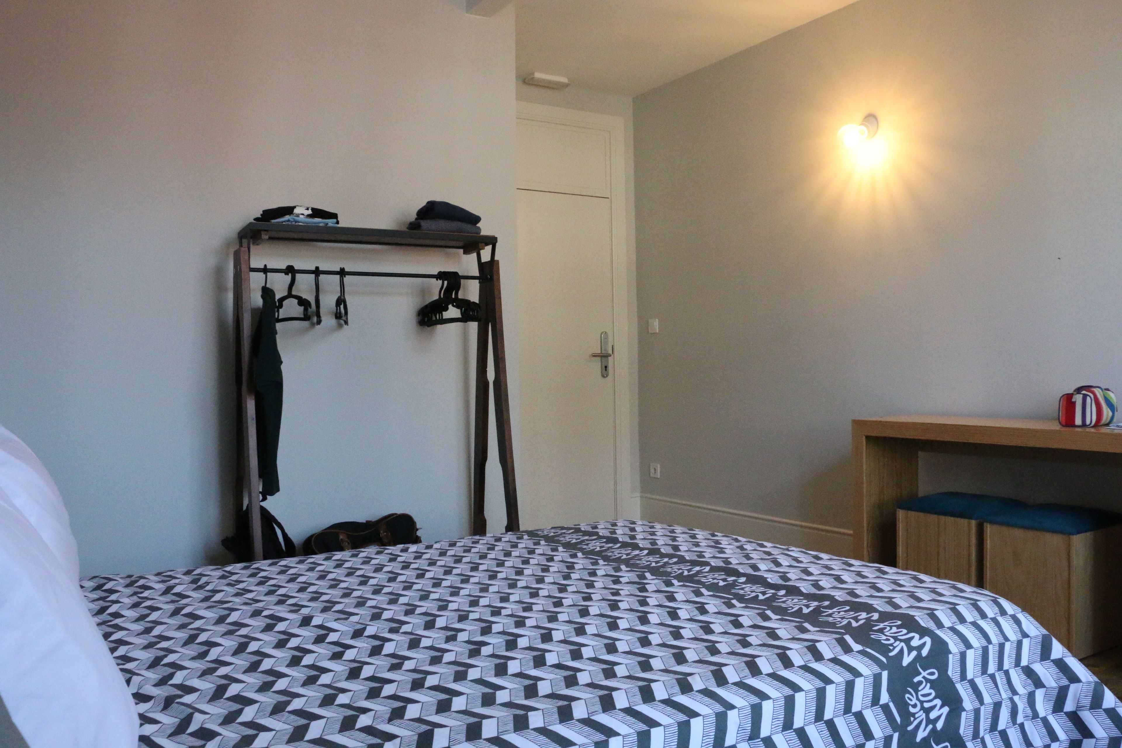 Quarto com cama, um cabideiro, uma porta e uma mesinha de cabeceira.