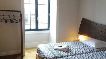 A hotel room with a bed, a window, a wall-mounted coat rack, and a bedside lamp.