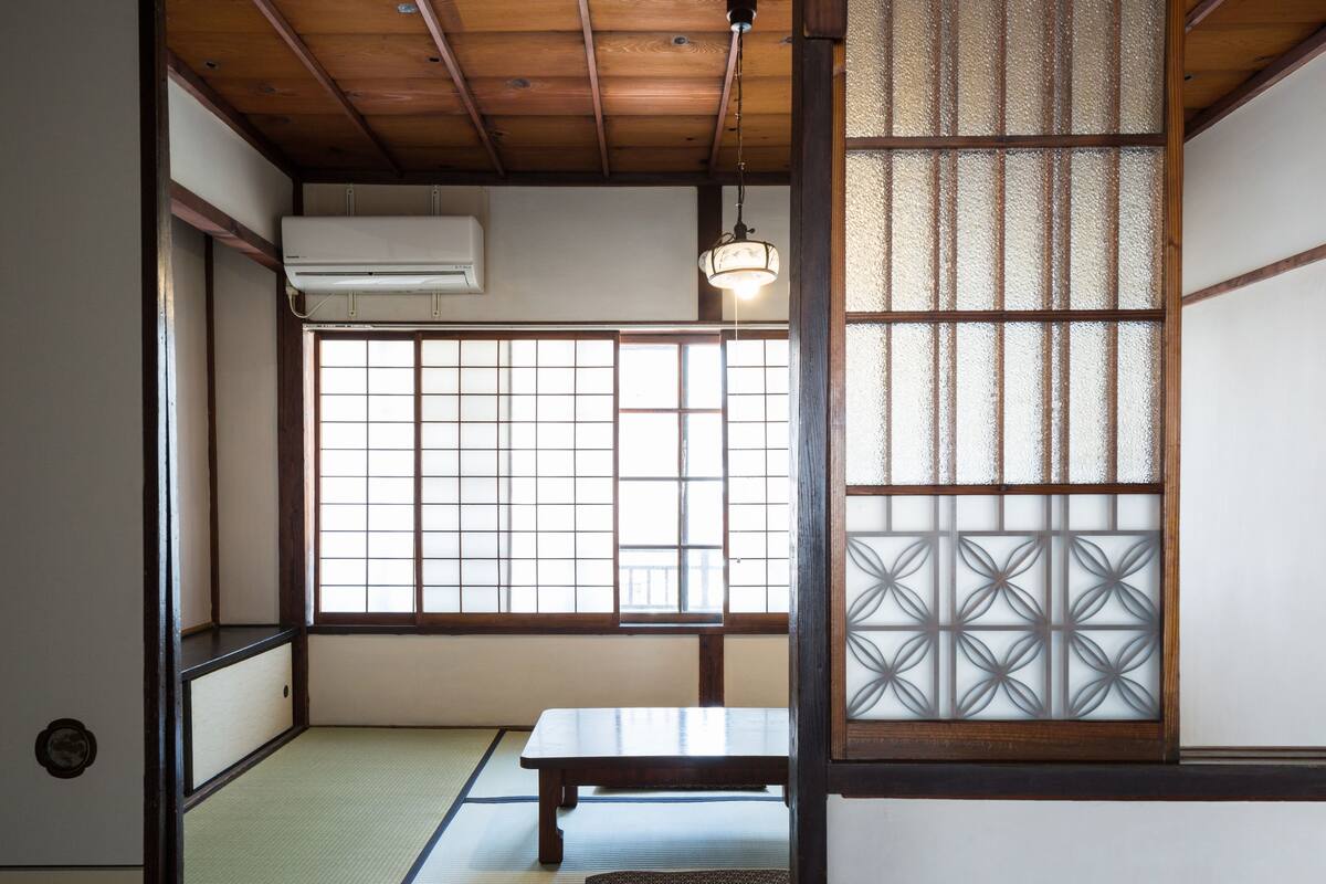 A traditional Japanese room with tatami flooring, a sliding door, a wooden table, and a hanging light fixture.