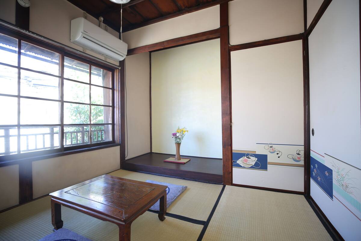 A traditional Japanese room with tatami flooring, a wooden table, a window with a view, and a sliding door.